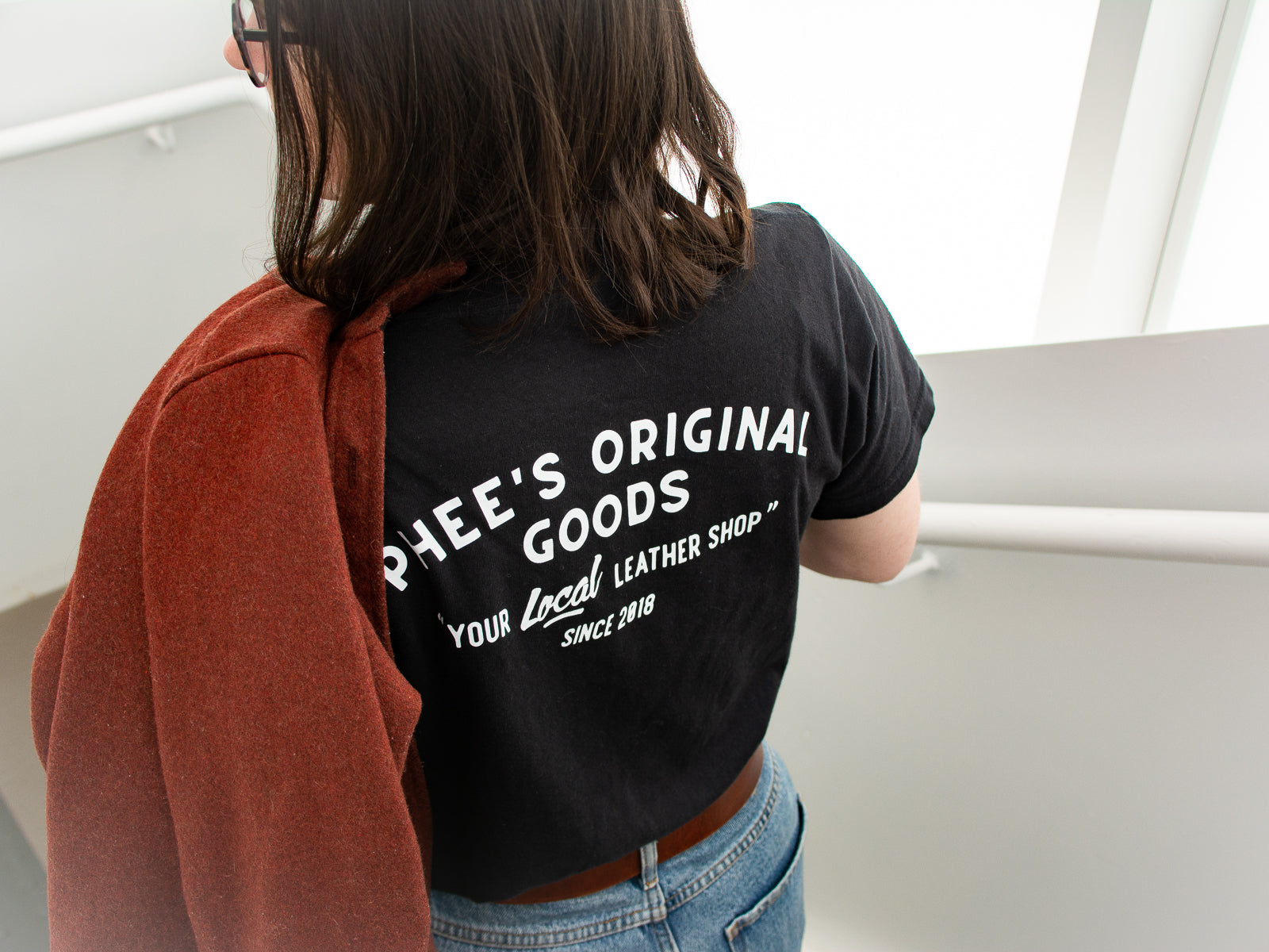 Robyn wearing a black anvil tee showing the "Your local leather shop" text while walking down a white staircase with a red sweater over her shoulder.