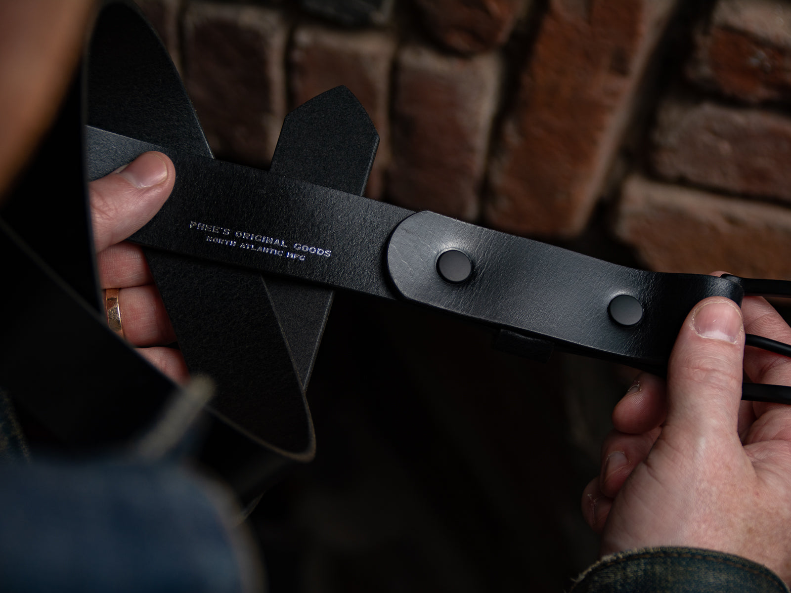 Person holding a black leather Ashfield belt, showing the inside with siver foil embossed logo and solid brass hardware.