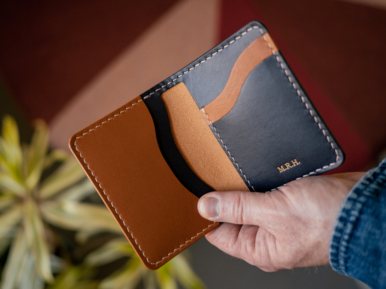 A hand holding a custom build your own barra wallet in navy and pine leather with taupe handstitching and gold embossing monogram