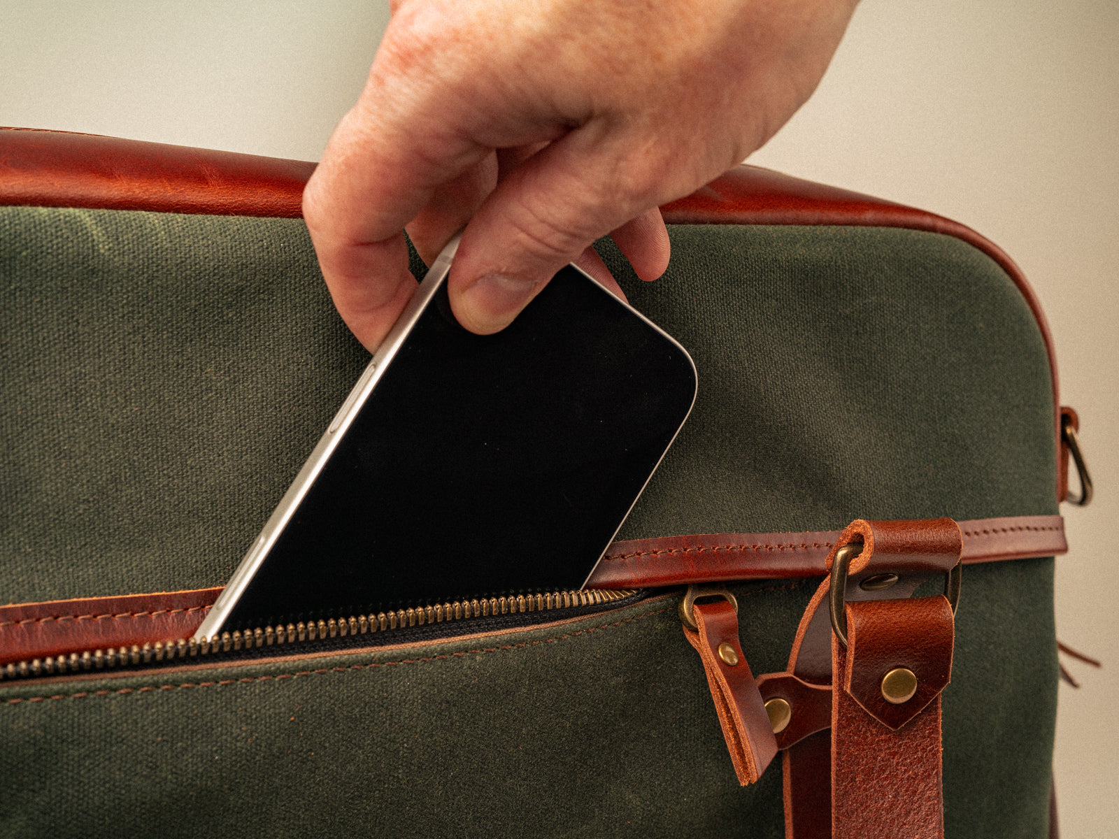 hand pulling a phone out of a hidden pocket on the back of the campbellton messenger bag in olive canvas