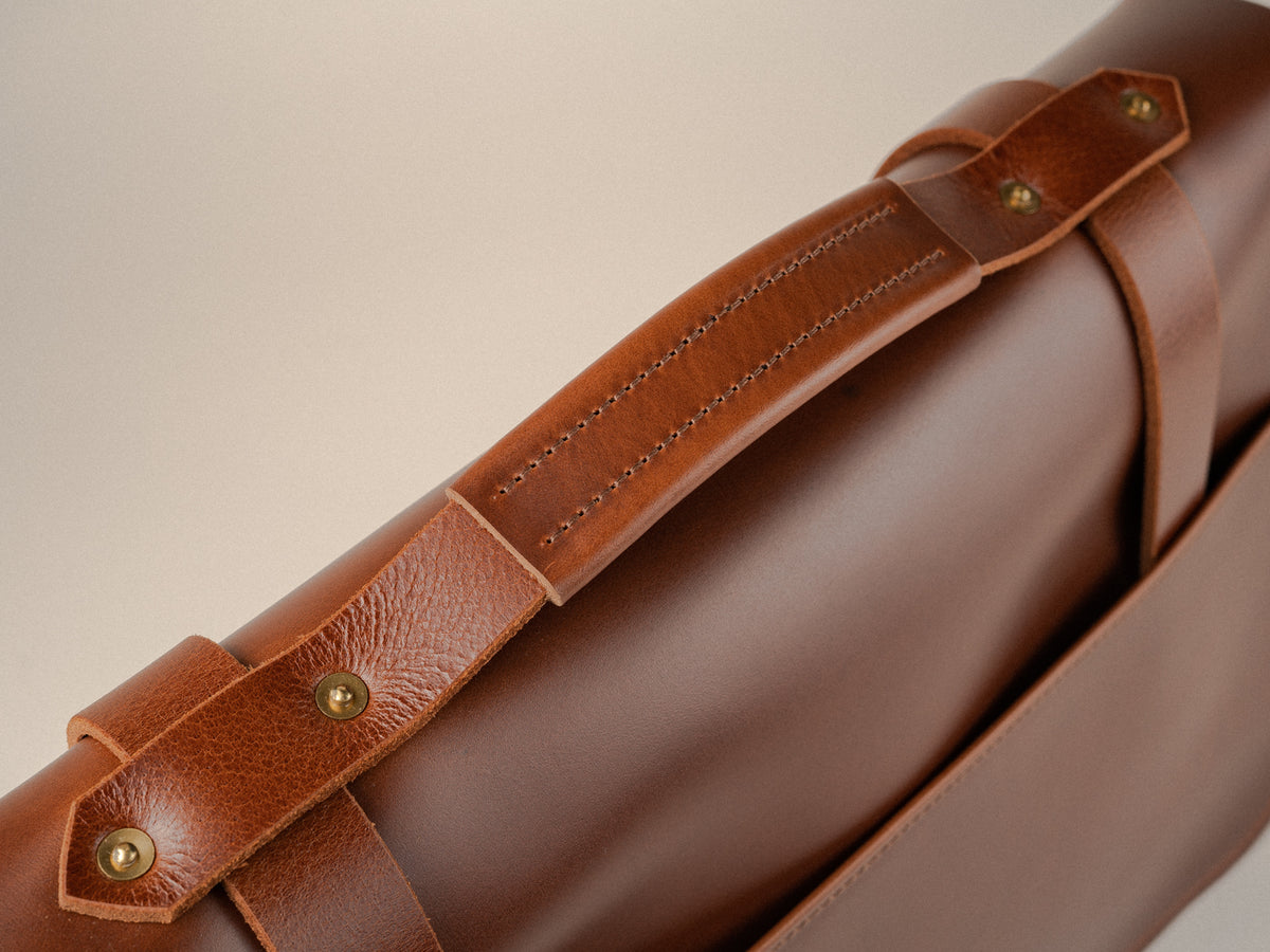 Chestnut full-grain leather messenger bag on a beige background, showing padded handle