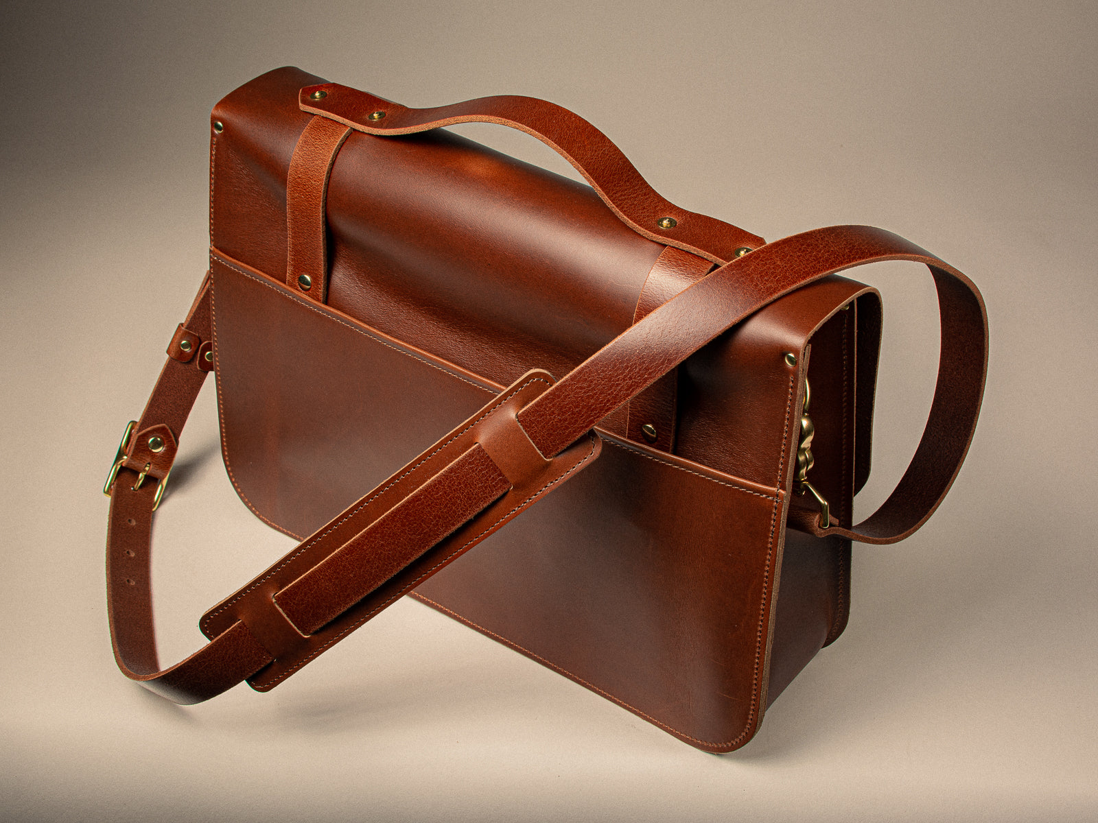 Chestnut full-grain leather messenger bag on a beige background, showing padded shoulder strap