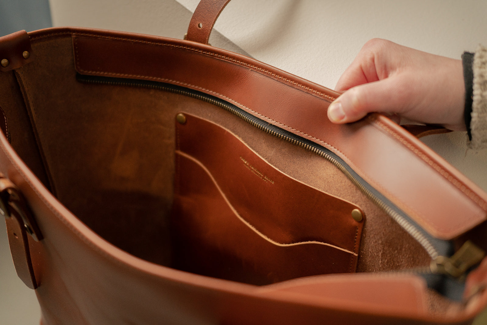 Chestnut tan Grand Etang leather bag with a hand opening it, featuring solid brass zipper and hardware.