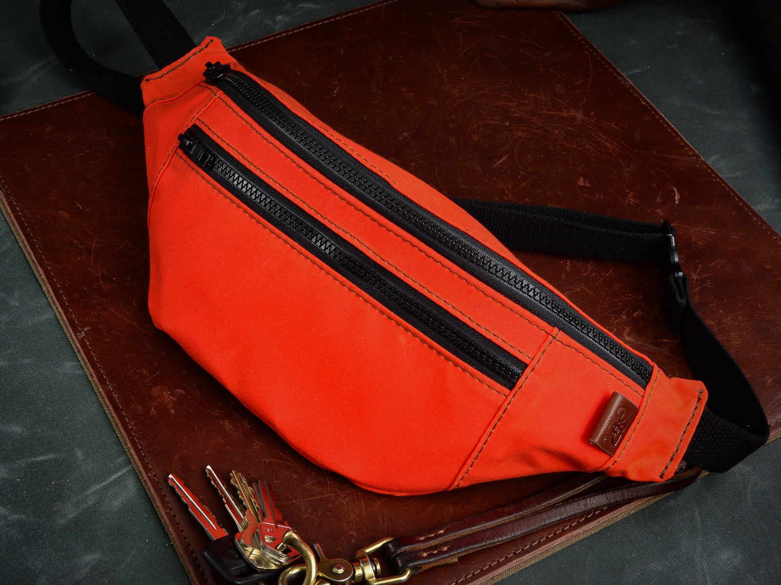 Hunter Orange Fanny Pack sitting on leather mat beside keychain with brown leather key lanyard. Fanny Pack has green stitchin, black zipper, black cotton webbing strap, and brown leather tag.