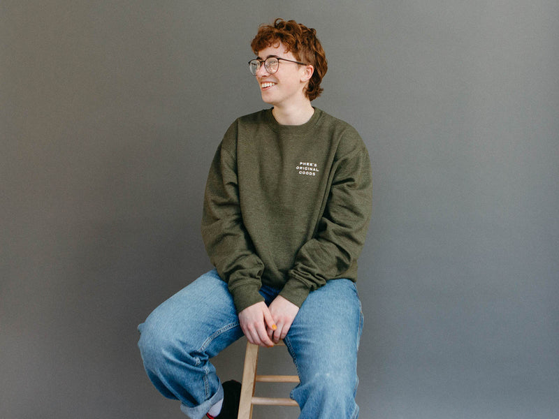 Mads sitting, wearing the heather olive Gull Crew Sweater, with Phee's Original Goods logo on the chest