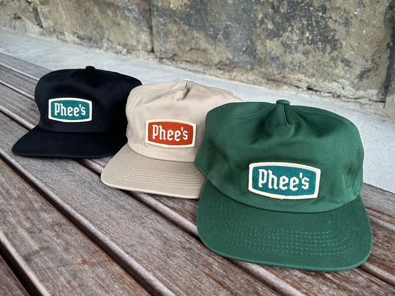 All three colours of the Phee's Badge Hat with embroidered felt patch sitting on a wooden bench
