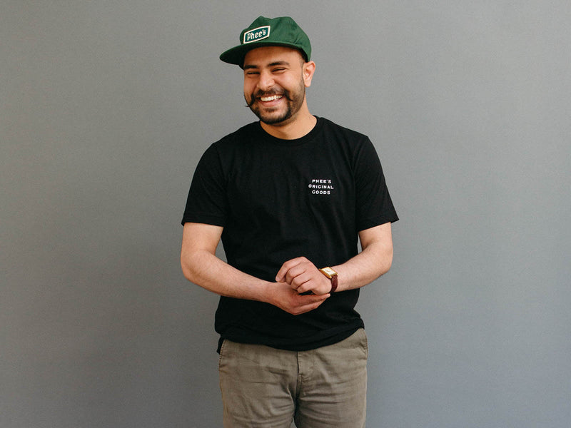 Shubham laughing, wearing the 100% cotton, sweatshop free Gull Tee Shirt in black, with Phee's Badge Hat in Pine