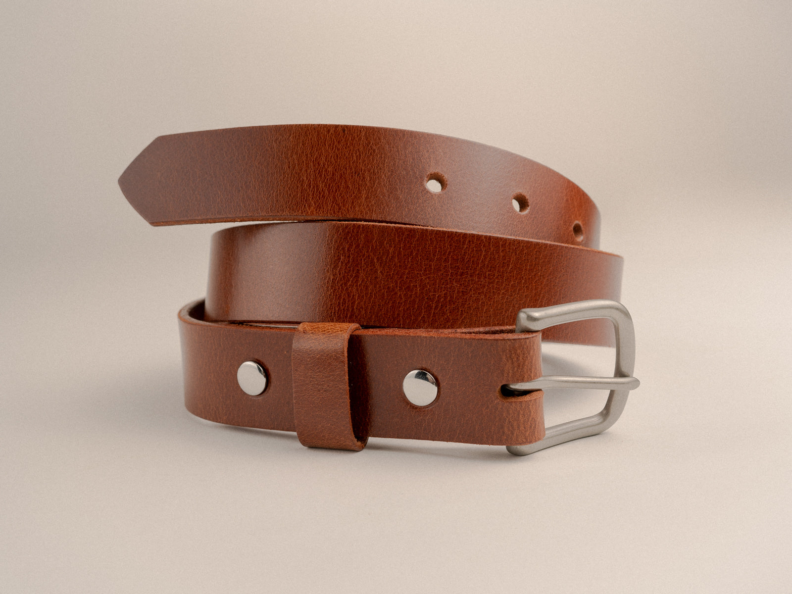 Warm tan brown leather belt with nickel finish solid brass hardware coiled on a white table