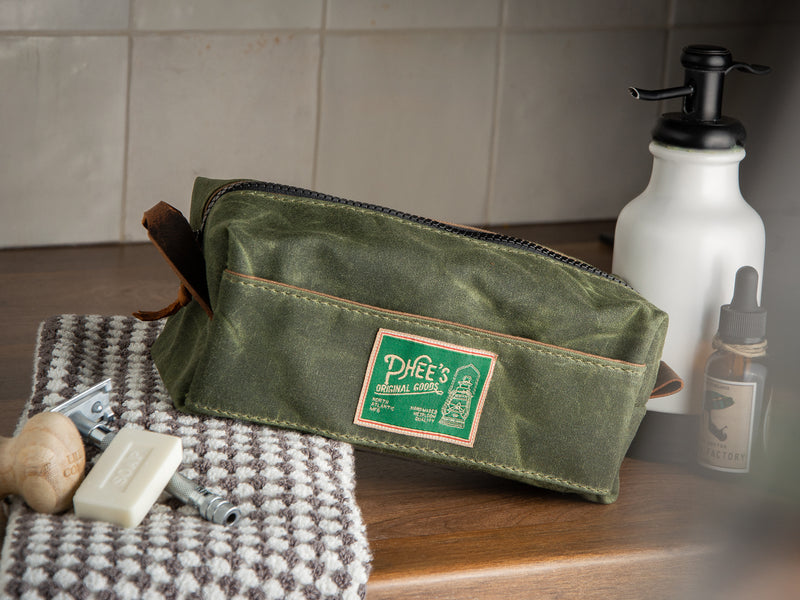 Olive green waxed cotton canvas Washabuck toilety bag sitting on a bathroom vanity wth shaving accessories