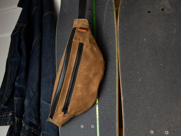 Handmade waxed canvas nutmeg Franey fanny pack hanging off of a cruiser skateboard