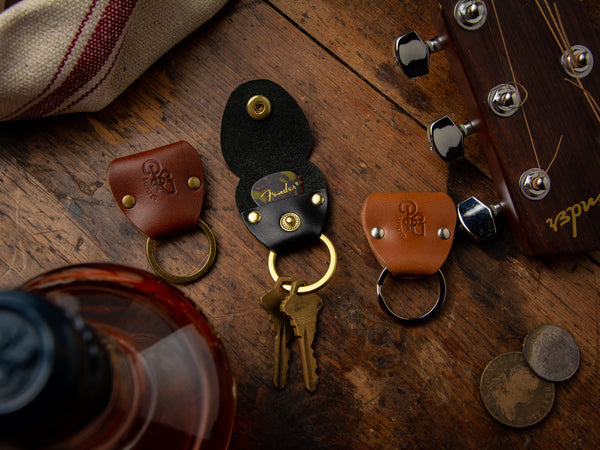 Guitar pick keychains in full grain leather laid out on a table beside an acoustic guitar