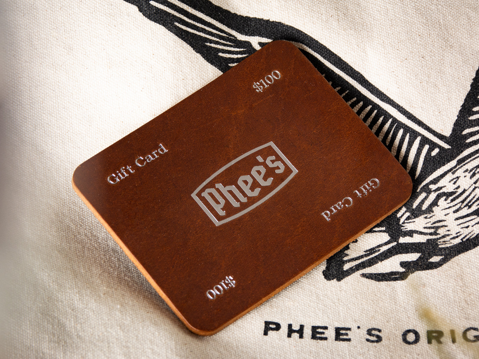 A leather gift card is pictured on top of a phee's original goods tote bag.