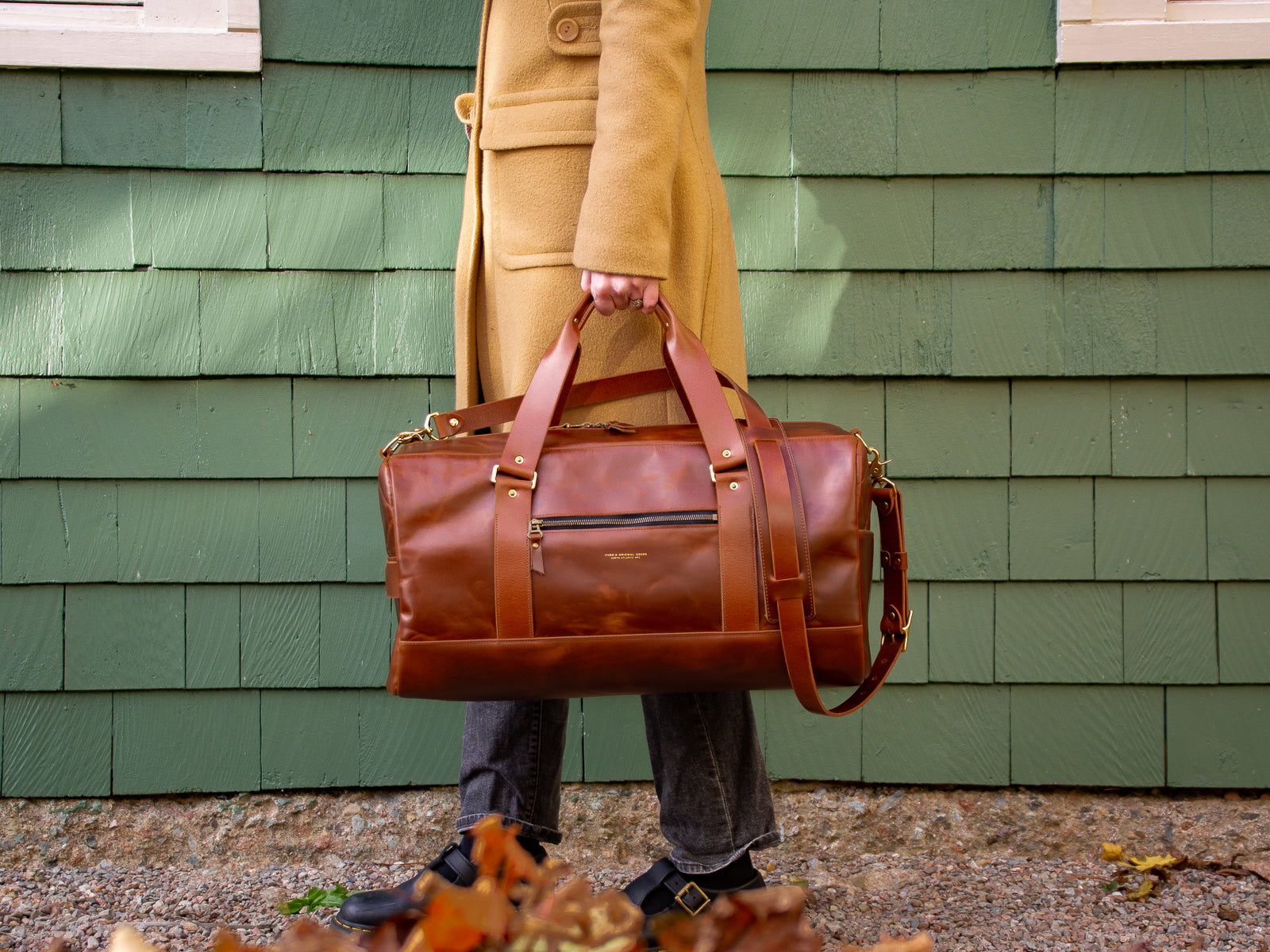 Reserve duffel bag in Chestnut Tan colour being held by a woman's side in the fall leaves.