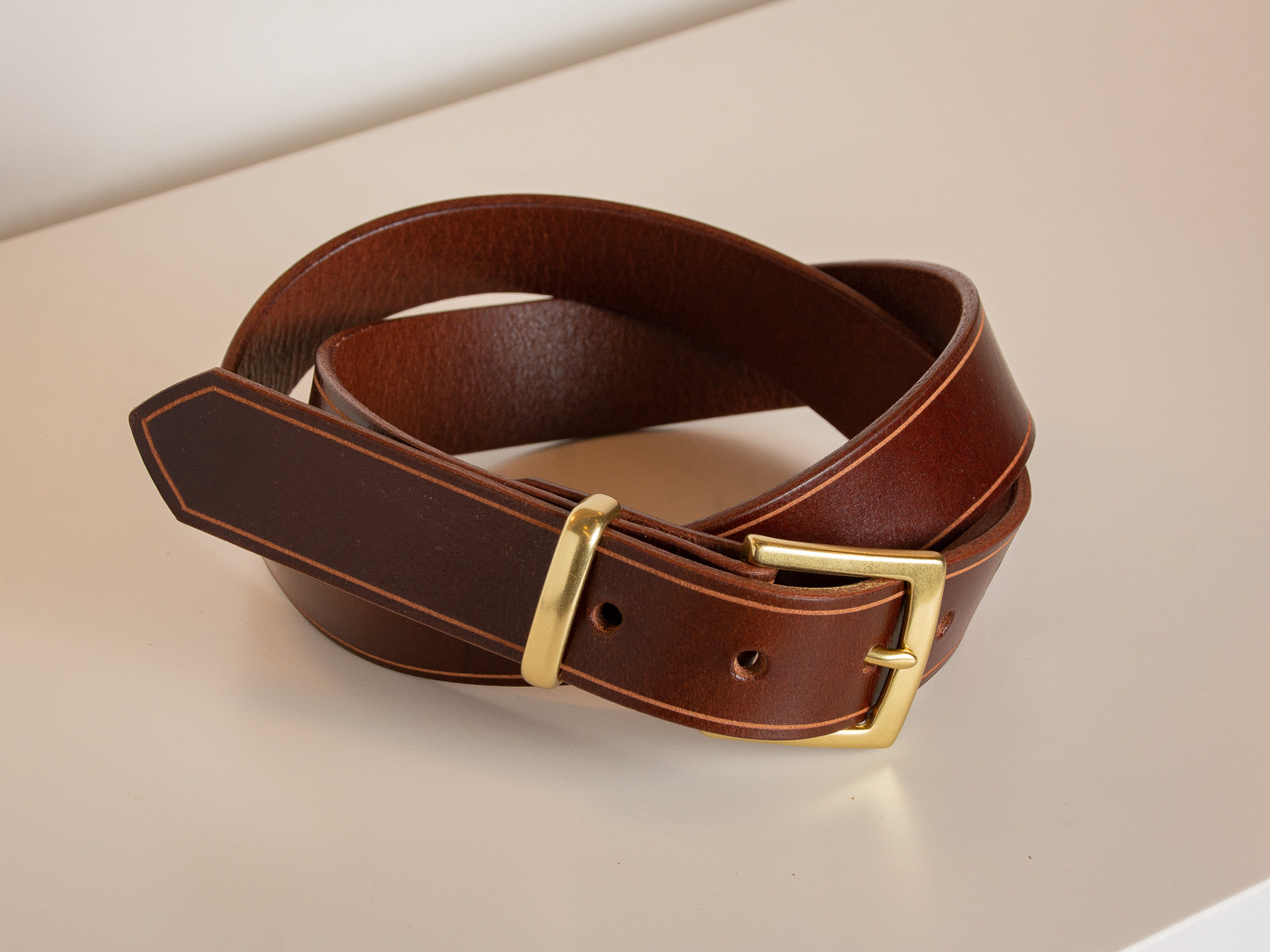 The Sterling Dress Belt in brown with natural brass buckle and keep on a white table.