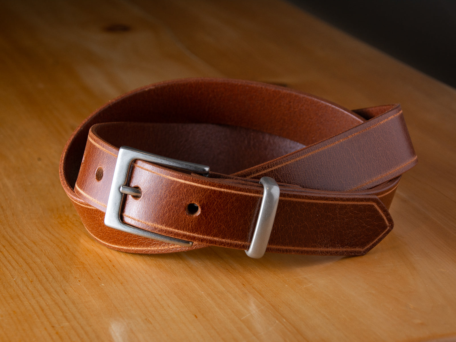 The Sterling Dress Belt in tan shown with nickel buckle and keep shown coiled on a wooden table.