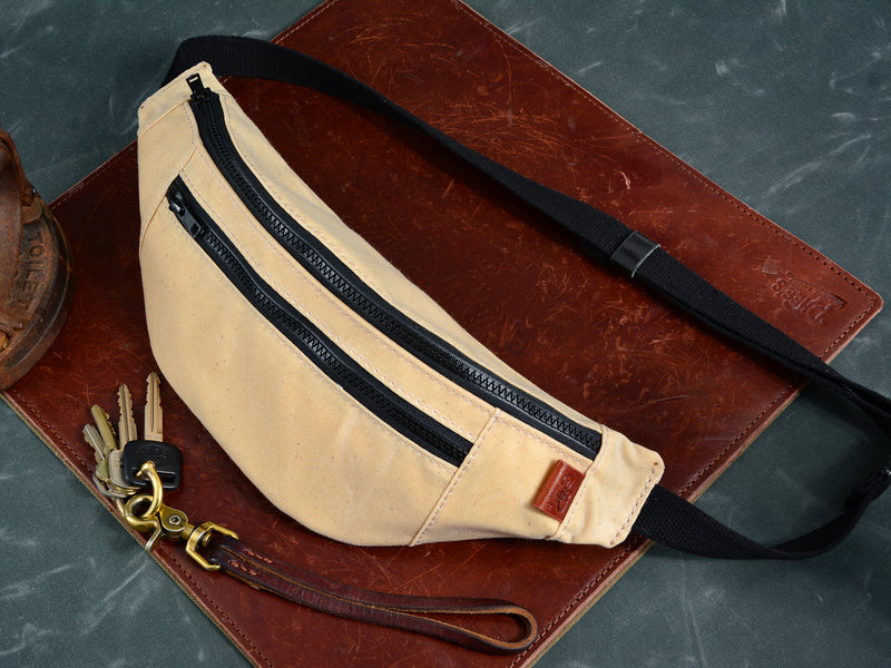 Cream coloured fanny pack sits on brown leather mat beside antique iron and brown leather key lanyard. Fanny pack has cream stitching, Black cotton webbing strap, and brown leather tag.