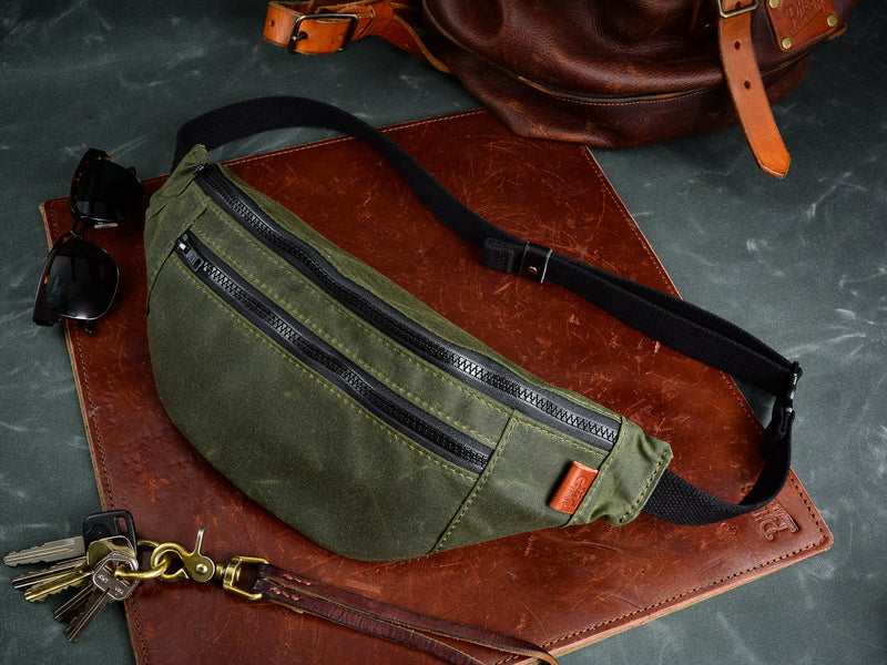 Green Fanny pack sits on brown leather mat beside sunglasses, key lanyard and bottom of leather backpack in background. The fanny pack features green stitching, black zipper, black cotton webbing strap, and brown leather tag.