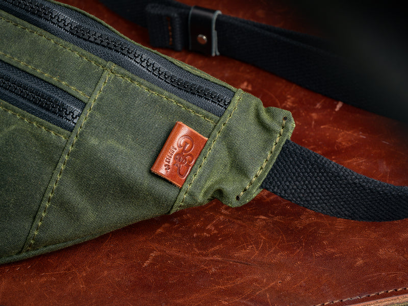 Close detail photo of black zipper, and brown leather tag on right side of green canvas fanny pack.
