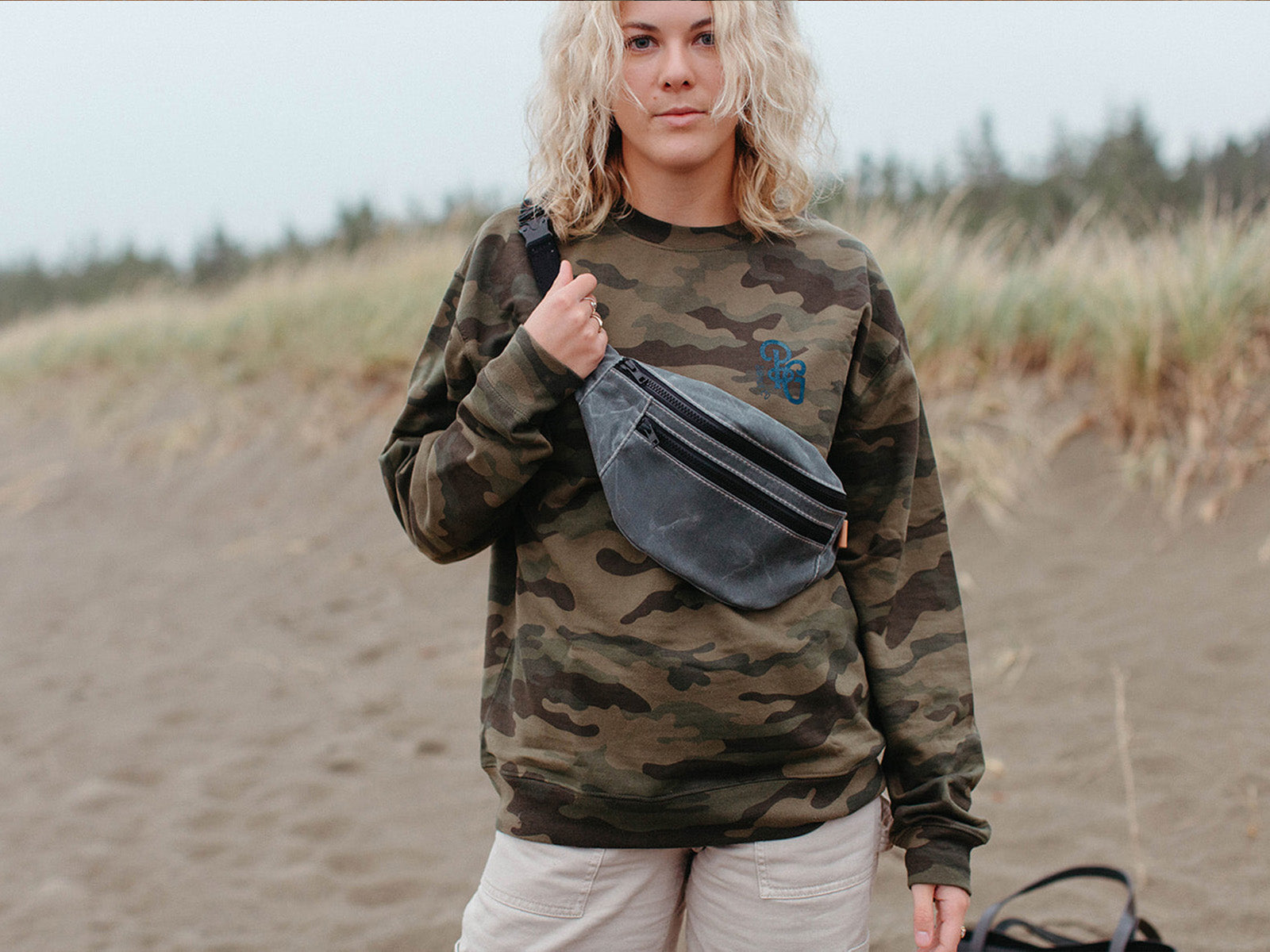 a slate grey waxed canvas fanny pack with black zip worn over a camo sweater at the beach