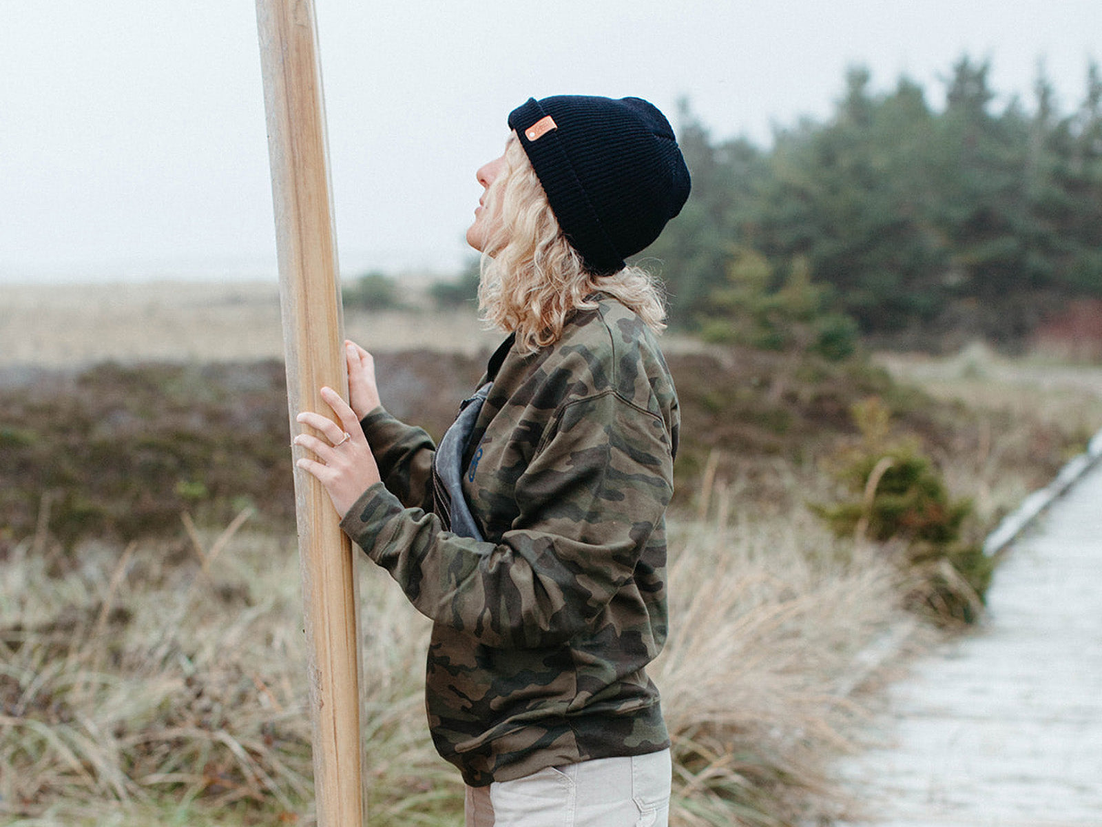 Nicole holding and looking up at her surfboard, wearing a black supply logo toque