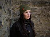 Robyn wearing a supply beanie in Olive, with a brown leather patch standing in front of a stone wall