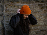 Robyn fixing her orange supply toque hat with brown leather patch, standing in front of a stone building