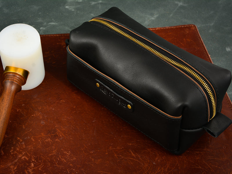 Black washabuck toiletry bag with brass zipper and hardware sitting on brown leather mat beside wooden mallet. 
